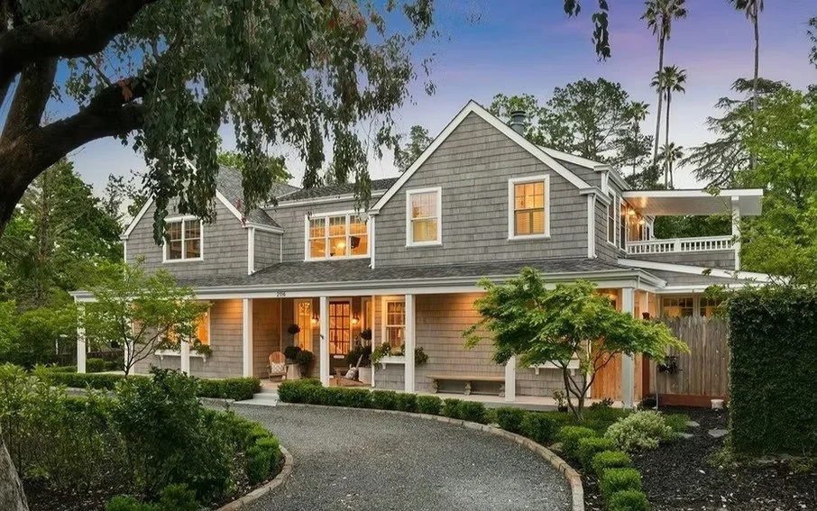 A large two-story house with grey shingles, white trim, and a porch, surrounded by greenery and tall trees.