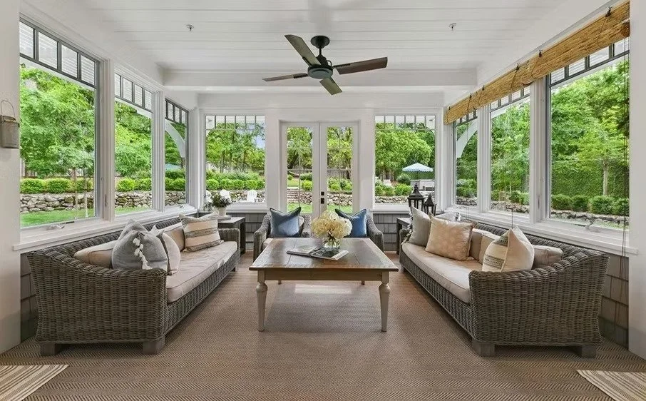 Sunroom with two wicker sofas with beige and gray pillows, a wooden coffee table with a flower arrangement, large windows with views of greenery, ceiling fan, and a door leading outside.