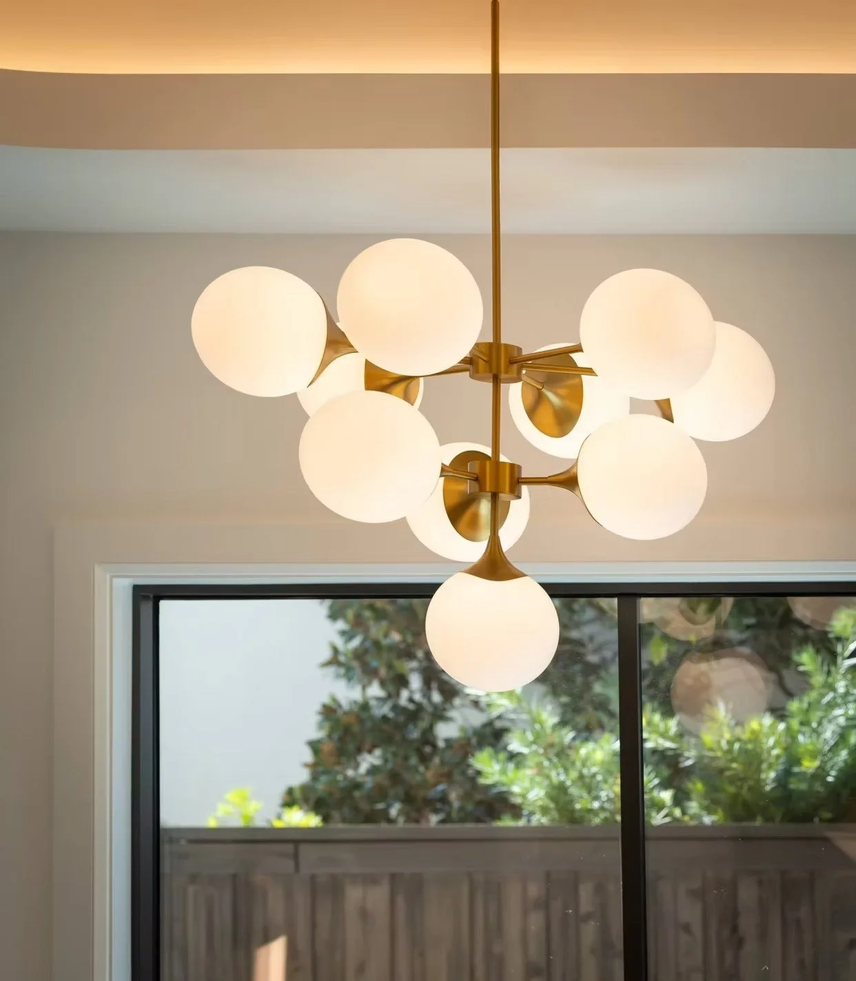 Modern chandelier with multiple white globe lights hanging from a central gold structure, inside a room near a window with greenery outside.