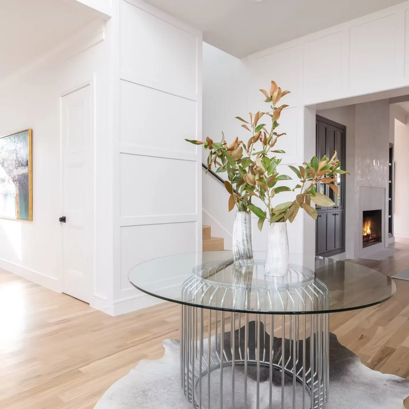 Interior of a modern living space with white walls, a glass coffee table, two vases with large leafy branches, and a fireplace in the background.