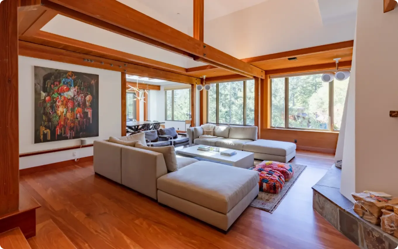 Living room with large sectional sofa, wooden beams, large windows showing trees outside, contemporary wall art, and hardwood floors.