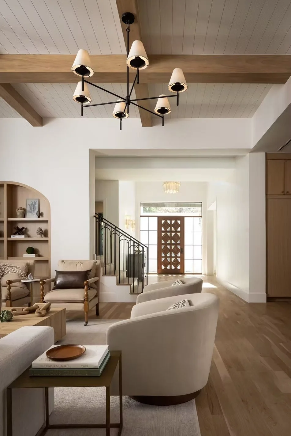 Living room with beige armchairs, a wooden side table with books and a tray, a built-in shelf with decorative items, wooden floors, a modern chandelier, and a staircase leading to the front door with large windows letting in natural light.