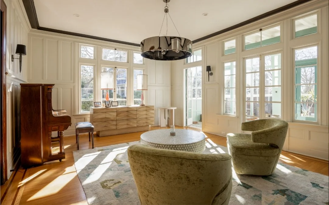 Bright living room with large windows, beige armchairs, a round ottoman, and a wooden sideboard. Sunlight streams through the windows, and a modern chandelier hangs from the ceiling.