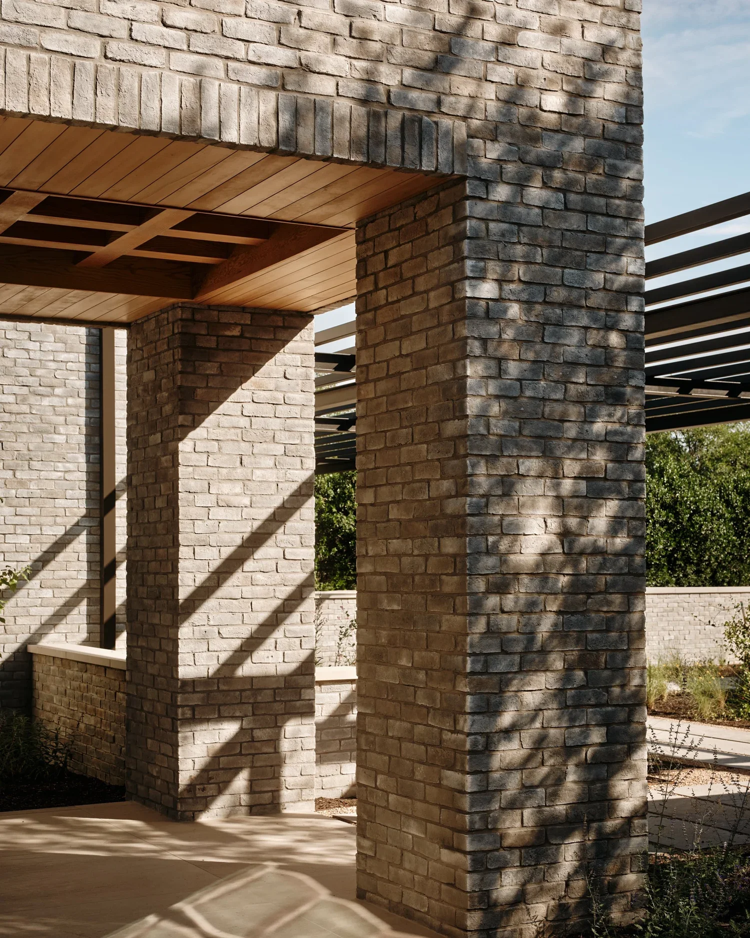 The image shows a modern building with brick columns, a wooden ceiling with an open grid structure, and a black metal railing on the right side. There are shadows cast by the eaves, and some greenery is visible in the background.