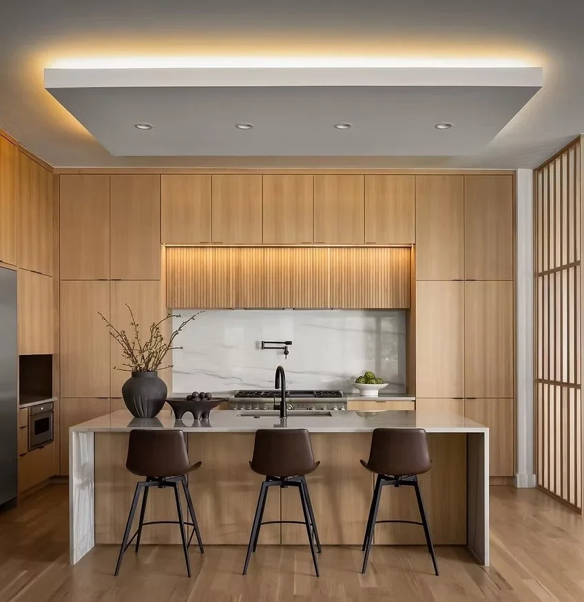 Modern kitchen with light wood cabinets, white marble backsplash, and a kitchen island with three dark brown chairs, decorated with a black vase, a bowl, and a bowl of fruit.