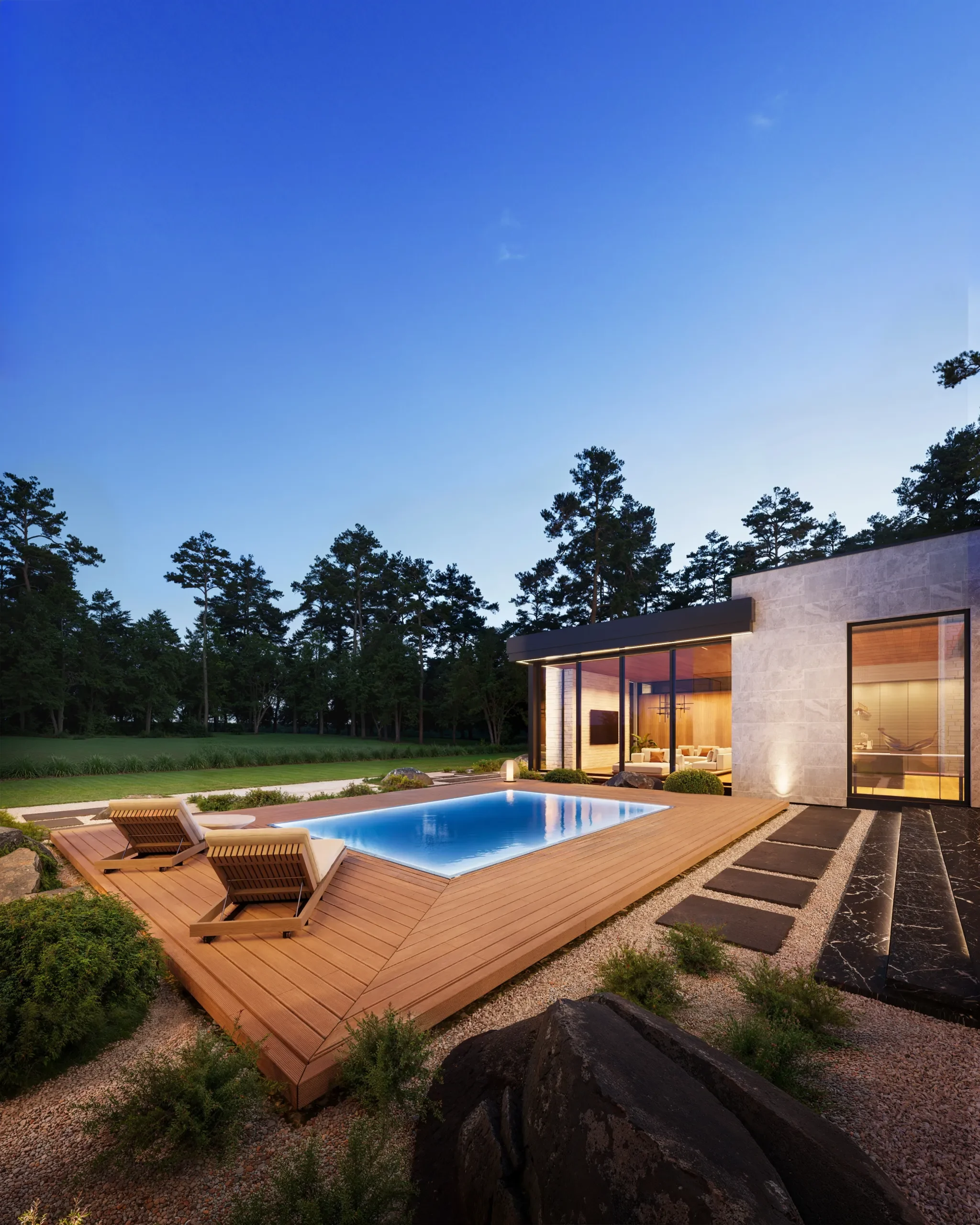 Modern house with large windows, outdoor swimming pool with wooden deck, lounge chairs, surrounded by trees and landscaped yard during dusk.