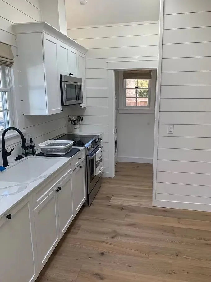 White kitchen with shiplap walls, black handles, stainless steel microwave and oven, white sink with black faucet, dish drainer, and small window with a view of a backyard.