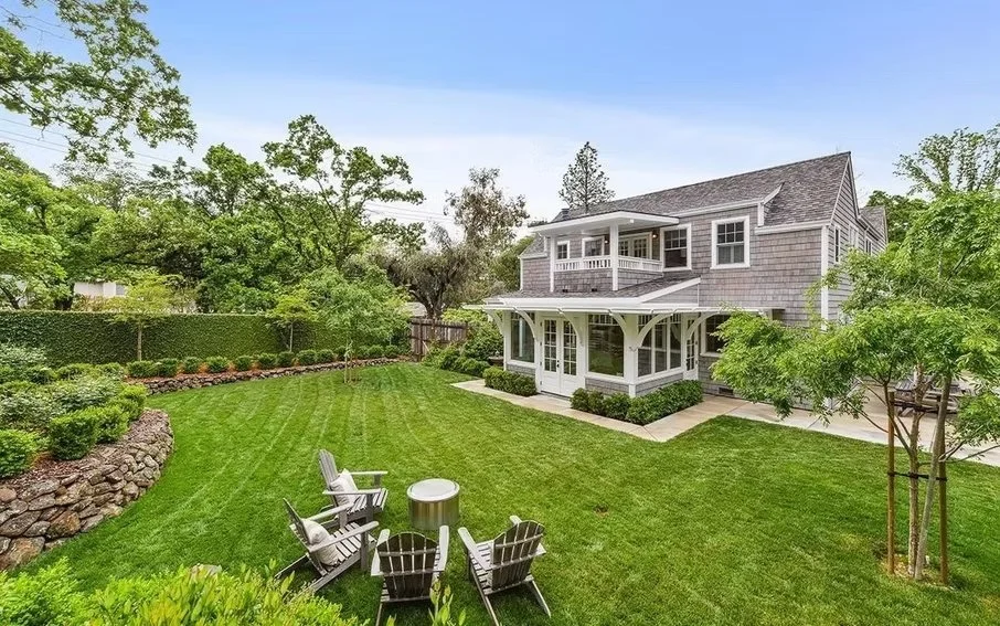 A spacious backyard with a neatly manicured lawn, surrounded by various trees and shrubs. There are four white Adirondack chairs arranged in a semi-circle around a small round table, overlooking the yard. In the background, a two-story house with gra