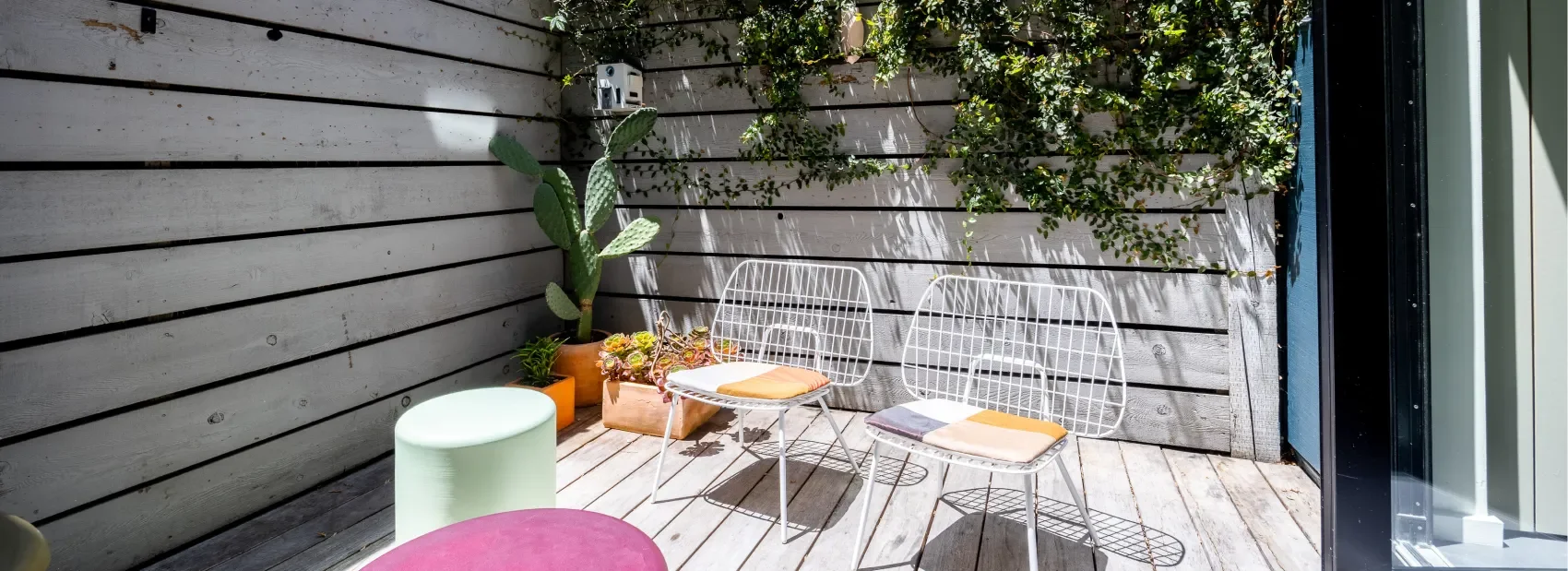 A small outdoor patio with wooden flooring, two white wire chairs with orange cushions, a green ottoman, a cactus plant in an orange pot, and a wooden planter box with succulents, against a wooden slat fence with green plants growing over it.
