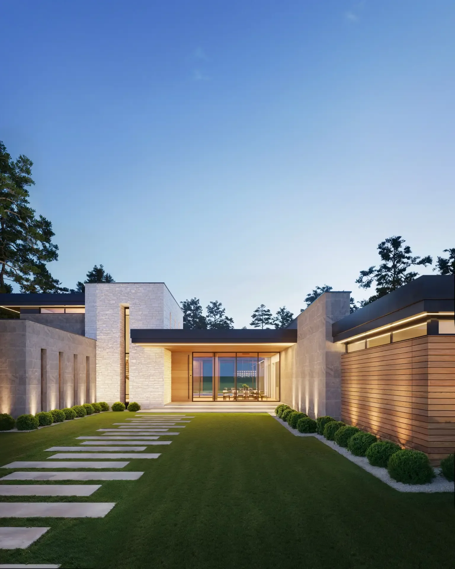 Modern house with a landscaped yard, illuminated exterior lighting, large glass doors, and a spacious patio area, surrounded by tall trees under a blue evening sky.