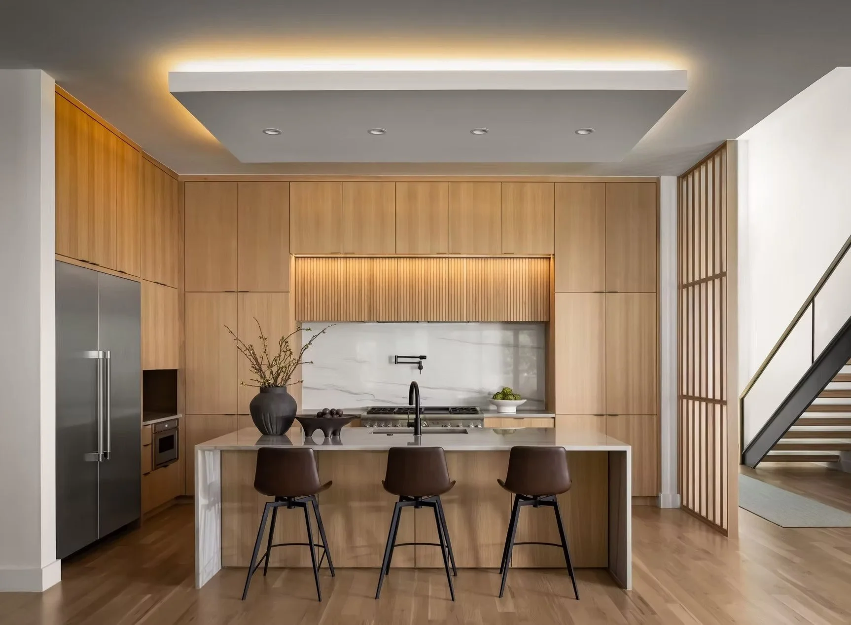 Modern kitchen with wooden cabinetry, marble backsplash, central island with seating, black faucet, and decorative items, including a vase with branches, placed on a chandelier, and a bowl of green apples.