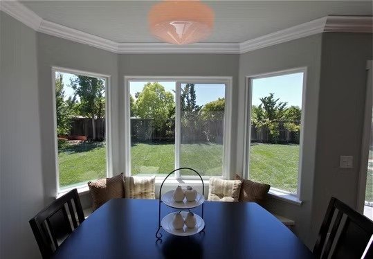 Dining room with a dark wood table, four chairs, and three pillows on window benches. Large windows showing a grassy yard with trees.