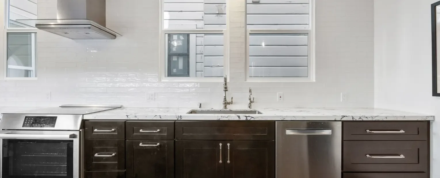 Modern kitchen with dark wood cabinets, white marble countertops, a stainless steel oven, dishwasher, and dual sinks under two windows with white shutters.