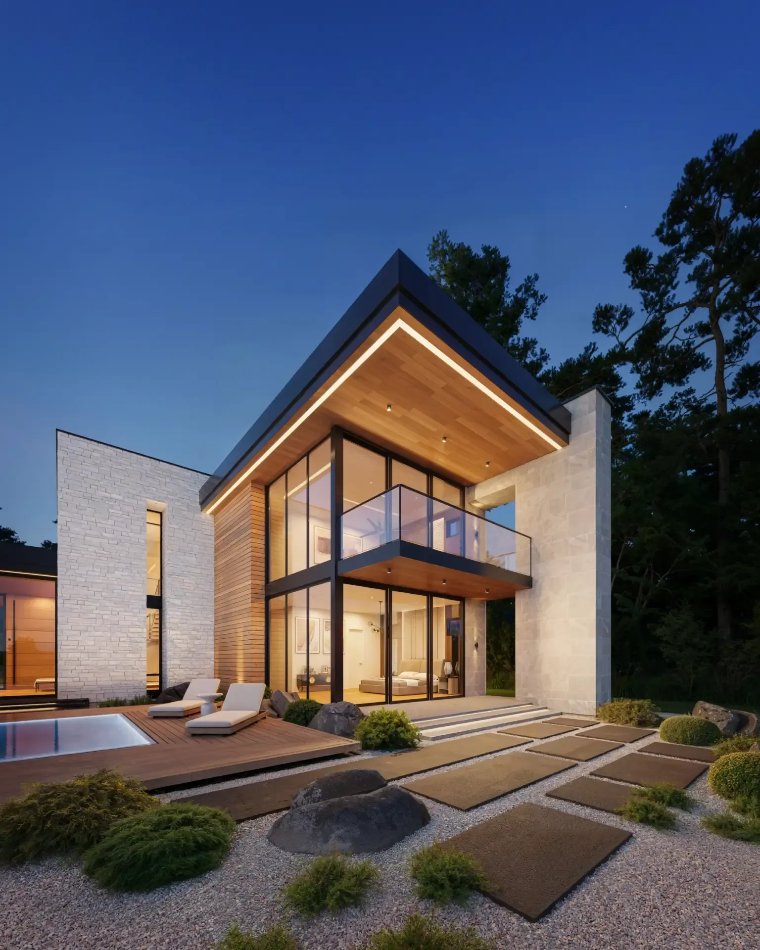 Modern two-story house with large glass windows and a balcony, surrounded by a landscaped yard with rocks, shrubs, paving stones, and outdoor lounge chairs, during twilight.