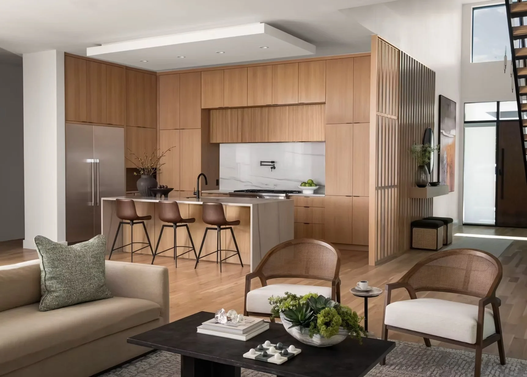 Modern open-concept living room and kitchen with wood cabinetry, a white marble backsplash, a kitchen island with bar stools, beige sofas, armchairs, and decorative plants.