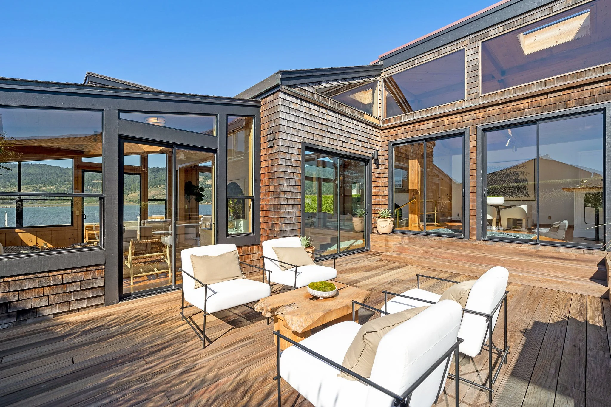 Modern house with large glass windows and wooden exterior, overlooking a lake and mountains, with outdoor seating on a wooden deck.