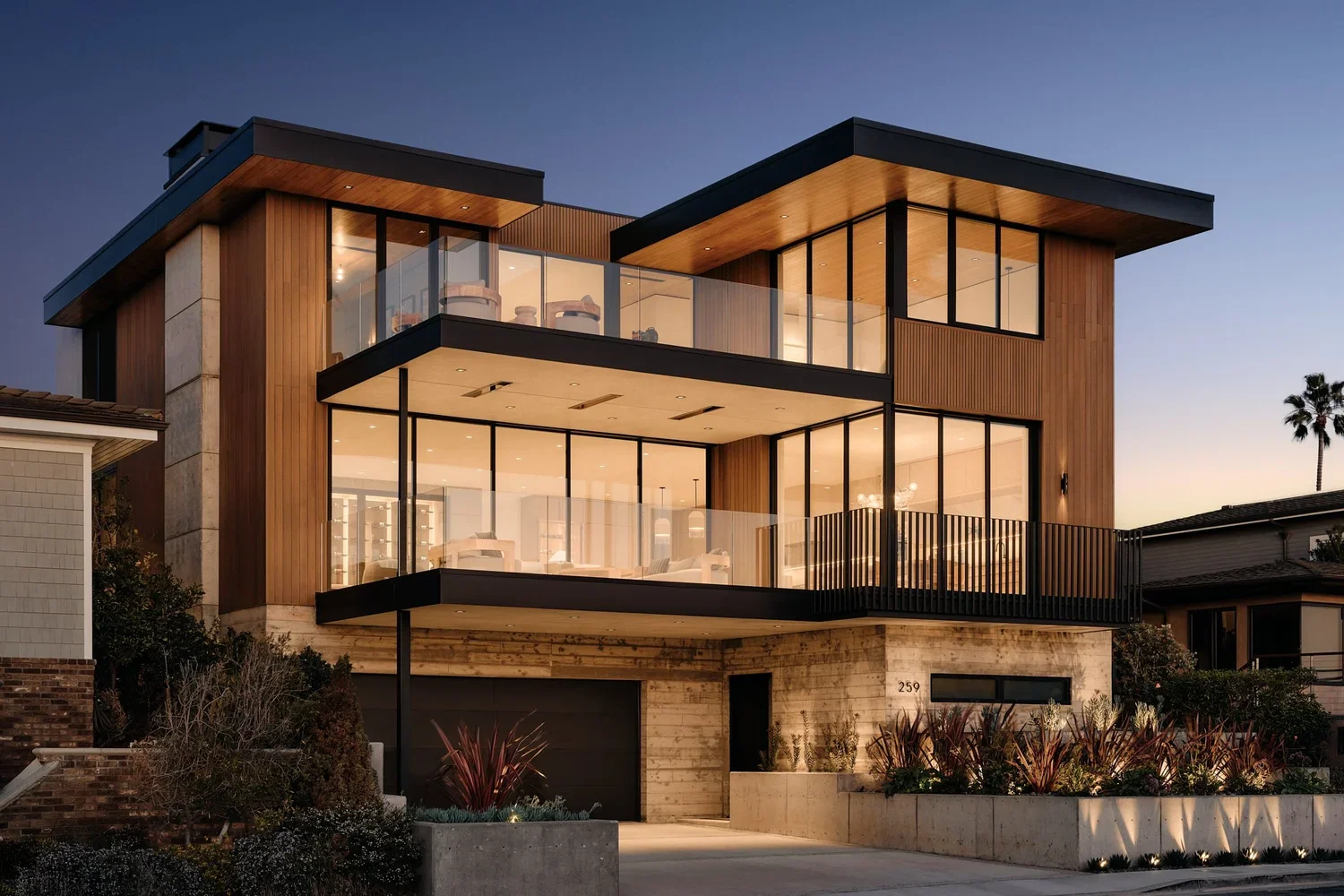 Modern three-story house with large glass windows, wood and concrete exterior, surrounded by landscaping, with palm trees in the background at dusk.