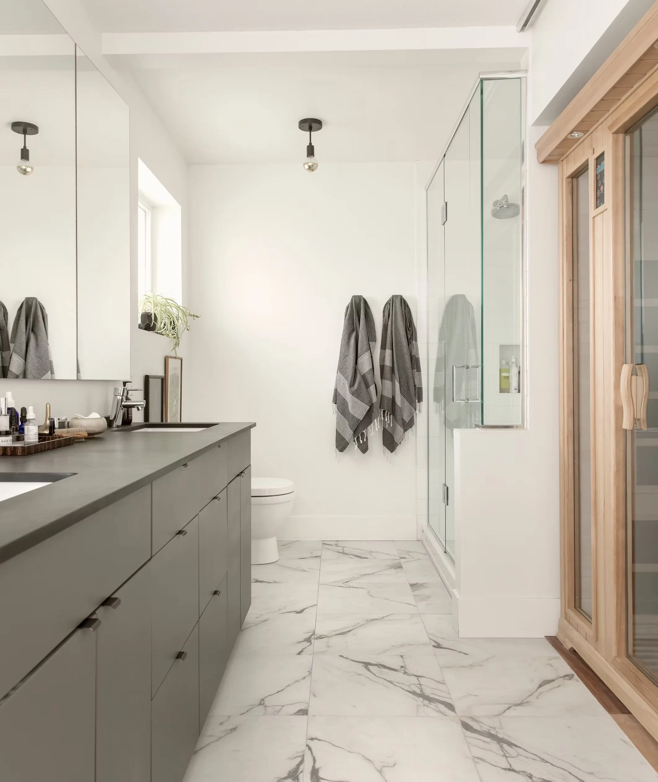 A modern bathroom with gray cabinets above the sink, marble flooring, a glass shower enclosure, a wooden sauna or cabinet on the right side, and towels hanging on hooks on the wall.