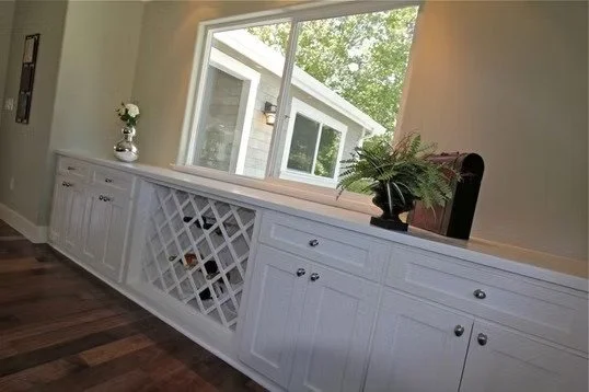 Sunlit room with white built-in cabinet, wine rack, potted fern, and vase with flowers, overlooking a backyard through large window.