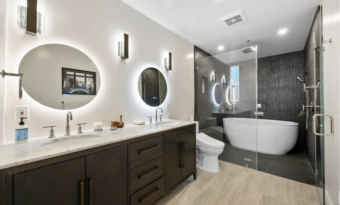 Modern bathroom with double vanity sinks, illuminated oval mirrors, bathtub, and shower area with glass partition, dark accent wall, and beige flooring.