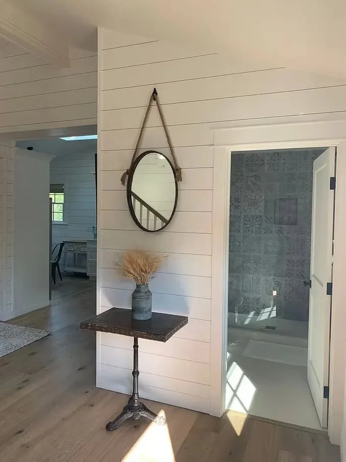 Interior of a modern home with a small table decorated with dried wheat, a mirror hanging on a white shiplap wall, and a doorway leading to a bathroom with patterned tiles.