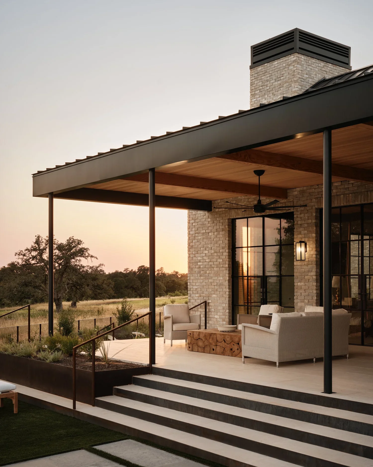 Modern outdoor patio with seating area, white chairs, and a wooden coffee table, under a black metal roof with wood paneling, brick wall, ceiling fan, and large windows, overlooking a natural landscape at sunset.