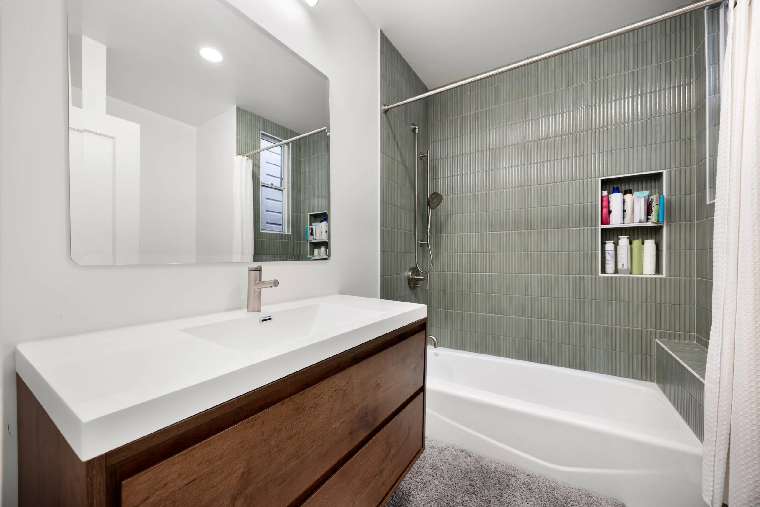 A modern bathroom with a white countertop, wooden vanity, large mirror, green tiled shower area with built-in shelves, and a small window.
