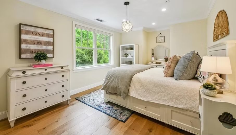 Bright bedroom with a white bed, decorative pillows, a nightstand with a lamp, a dresser with a pink tray, a window with lush green trees outside, and wall decor including a sign with 'LOVE YOU MADLY' and a small wall hanging.
