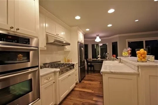 Modern kitchen with white cabinets, stainless steel appliances, granite countertops, and hardwood floors. There is a breakfast bar with a bowl of lemons, and a dining area with a window in the background.