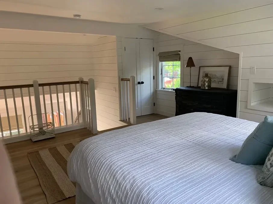 Bedroom with white shiplap walls, a bed with a white bedspread and pillows, a window with a blind, a small black dresser with a lamp and framed picture, and a railing overlooking a lower level.