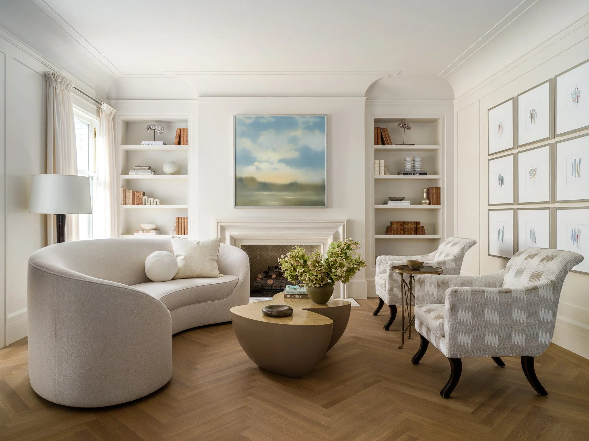 A living room with a curved white sofa, a wooden coffee table, an armchair with a checkered pattern, white built-in bookshelves, a large painting of a cloudy sky above a fireplace, and framed abstract art on the wall.