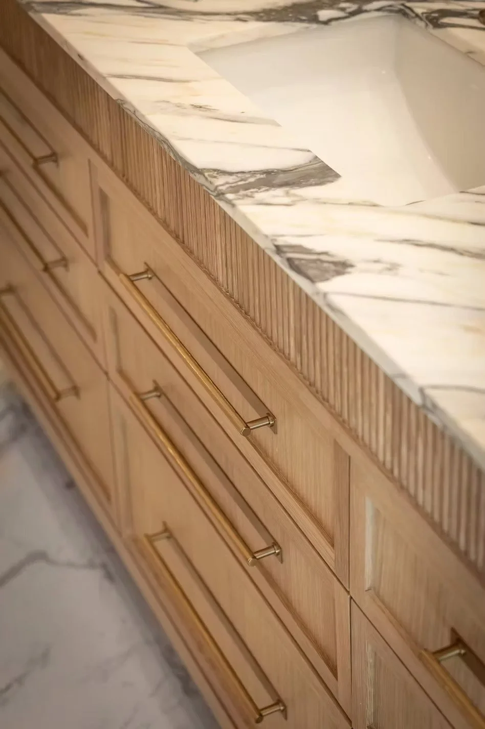 Close-up of a marble countertop with a built-in sink, wooden cabinet drawers with gold handles, and marble flooring.