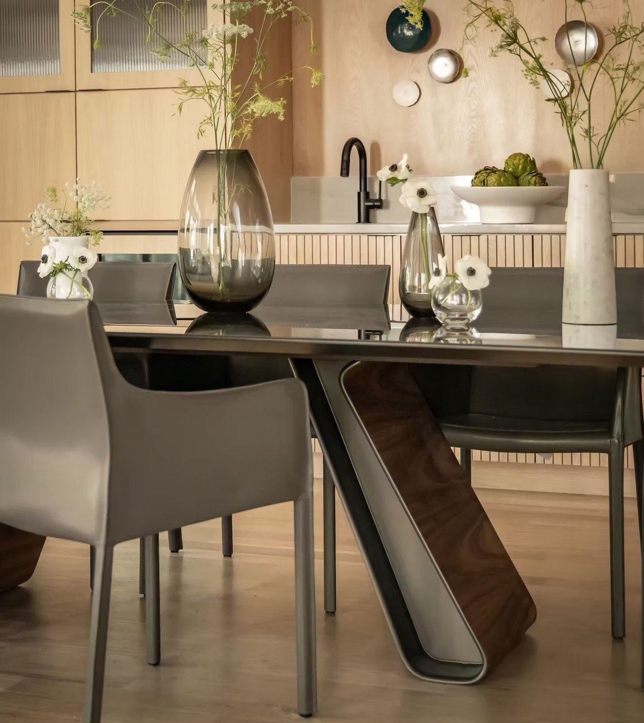 A modern dining room with a black rectangular table surrounded by beige and dark gray chairs. On the table are three decorative vases with white flowers and greenery. The background features a light wood kitchen counter with a black faucet and bowl o