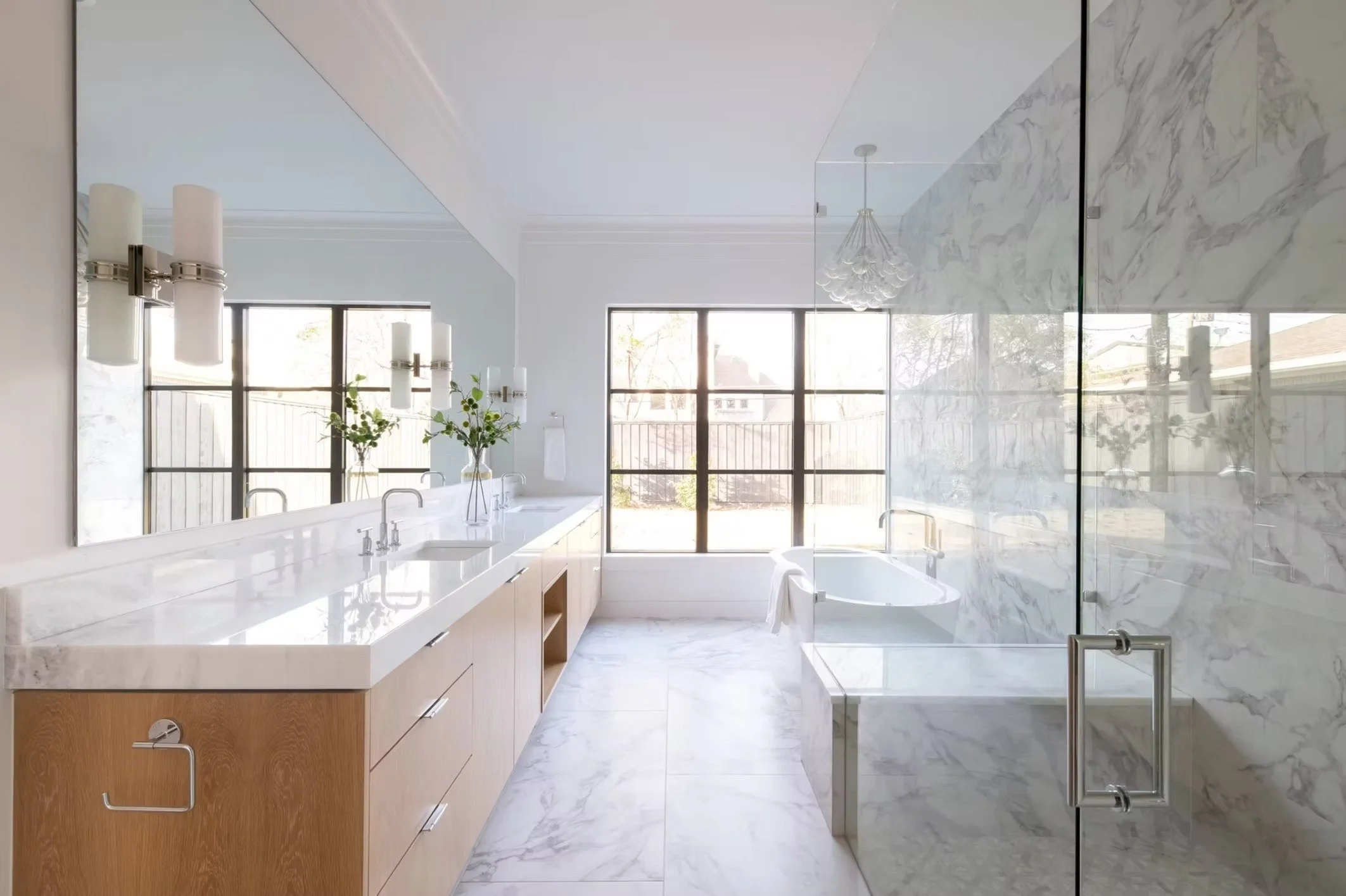 Modern bathroom with white marble tiles, a large mirror, and a wooden vanity with a white countertop. There are two sinks, glass window doors, and a bathtub with a towel hanging on the side. Decor includes green plants and unique light fixtures.