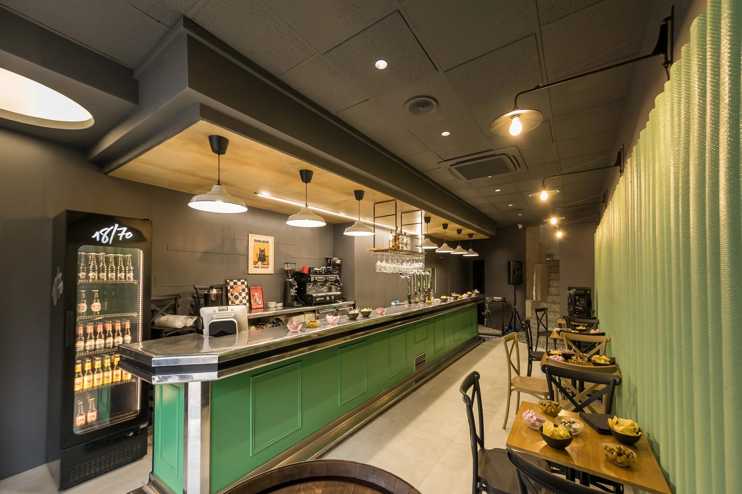 Interior de un restaurante con barra de bebidas verdes y mesas con sillas de madera. Hay bocadillos y snacks en las mesas pequeñas y luces colgantes sobre la barra.