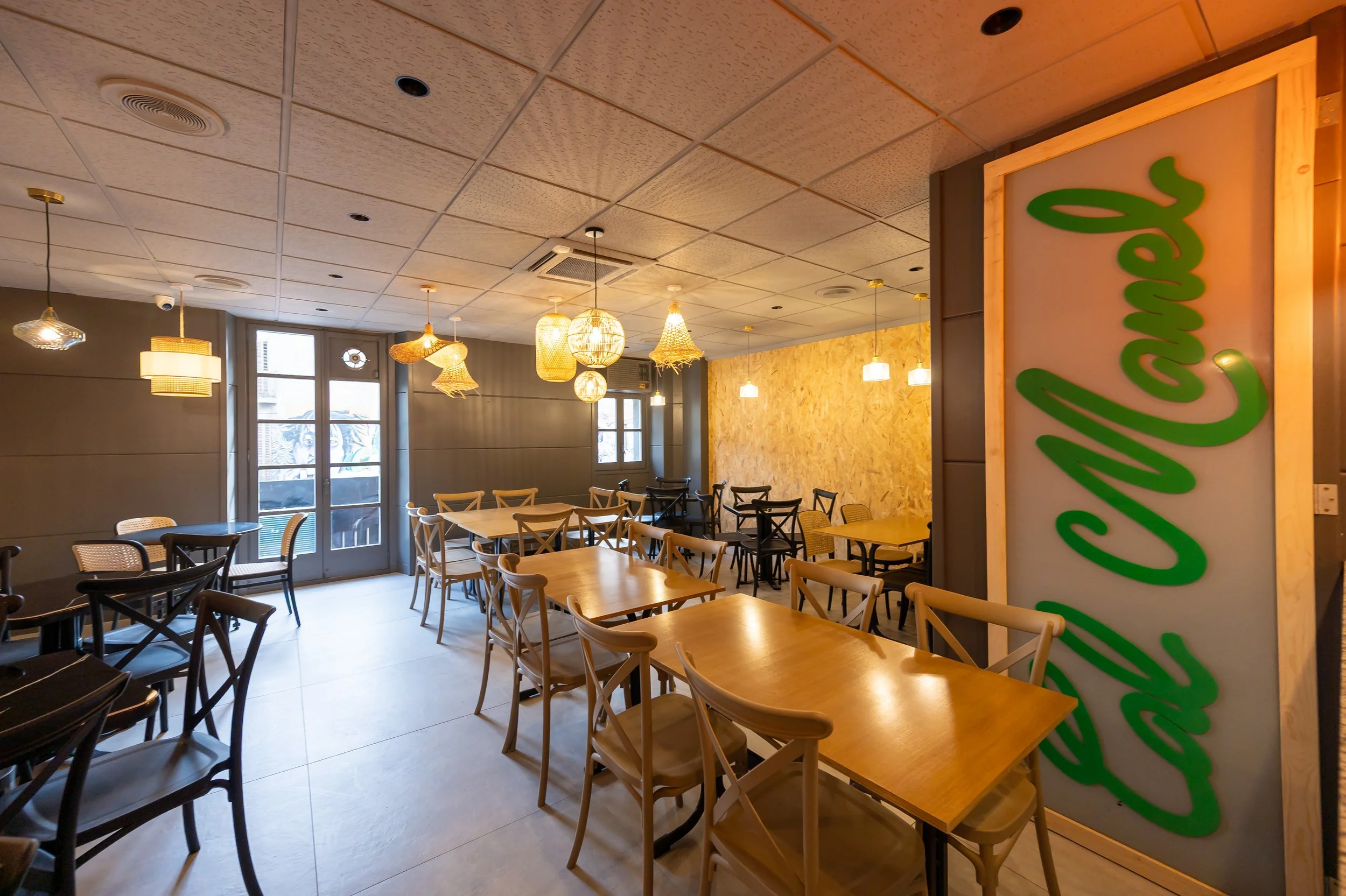Interior de un restaurante con mesas de madera y sillas, iluminación cálida y ventanas que dejan entrar luz natural.