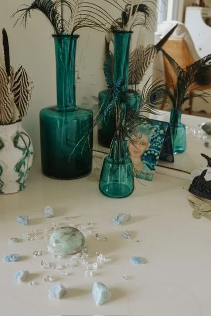 Arrangement of teal glass vases with feathers and greenery, a framed photograph, and decorative stones on a white surface.