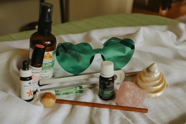 A collection of personal care items on a bed, including sunglasses, a spray bottle, a small dropper bottle, a roll-on perfume, a pink crystal, a large shell, and a wooden stick, with a white cloth background.