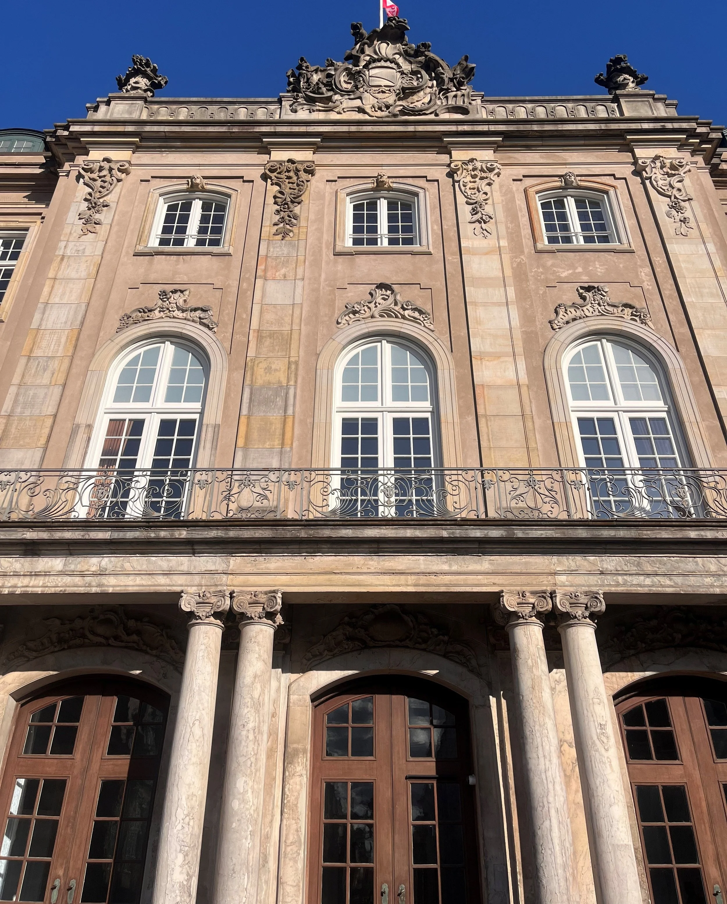 Front facade of an ornate historic building with arched windows, decorative stone carvings, columns, and wrought iron balcony railing, under a clear blue sky.