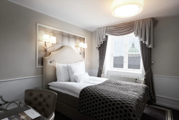 En soveværelsescene med seng, vindue med udsigt til bygningsdome, og moderne indretning.