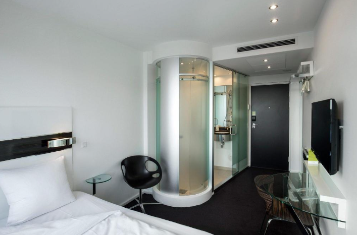 En hotelværelse med en seng, en sort stol, et glasbord, et fjernsyn og et mini-agtigt badeværelse med brusebad i en glasdør.