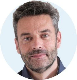 Et nærbillede af en mand med gråt hår og skæg, iført en mørk jakke, mod en lys baggrund.