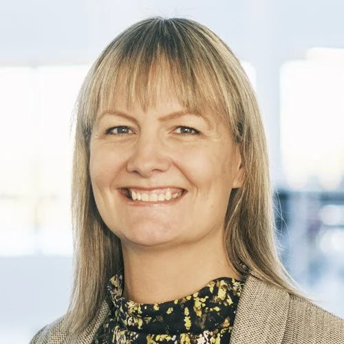 Kvindelig person med blondt hår, smilende og iført en mønstret top i en indendørsindstilling.