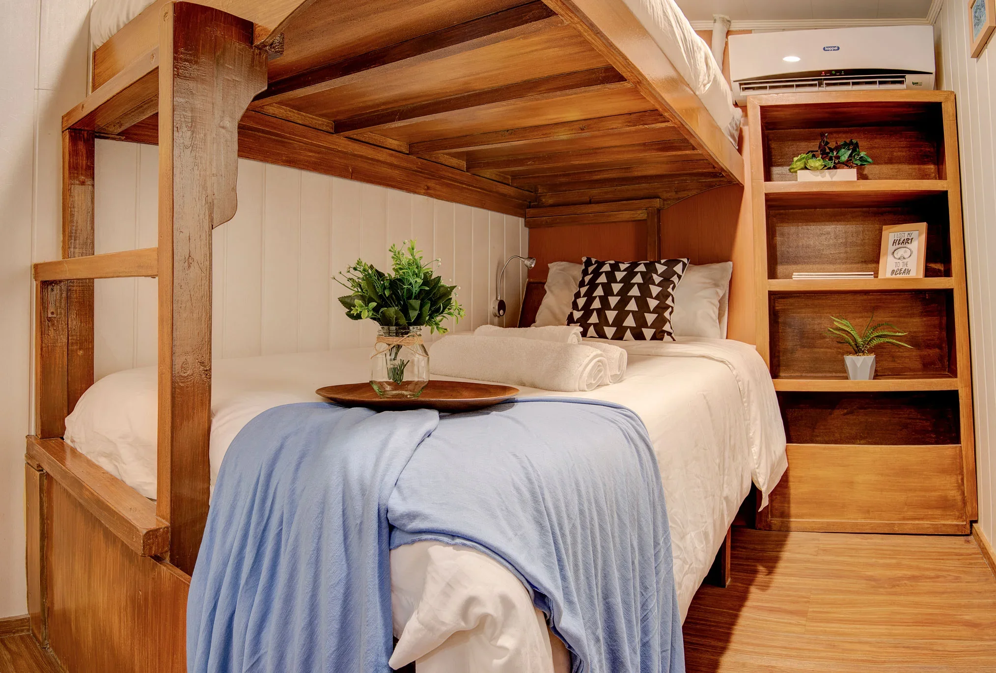 A cozy bedroom with a wooden bed frame, white bedding, a gray pillow with a geometric pattern, and a bedside table holding plants and towels. There's an air conditioner on the wall and hardwood floors.