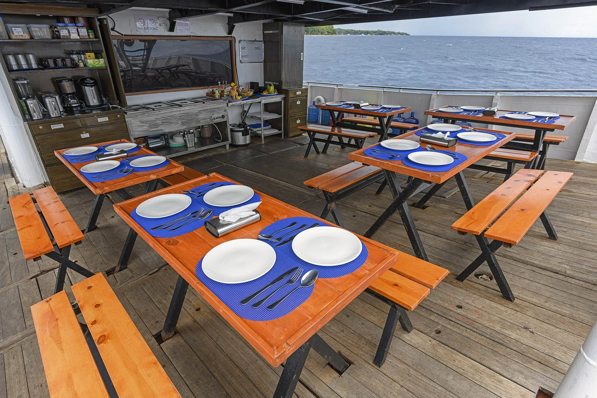 Outdoor dining area on a boat with wooden tables and benches, set with white plates, blue placemats, and silverware, overlooking water.