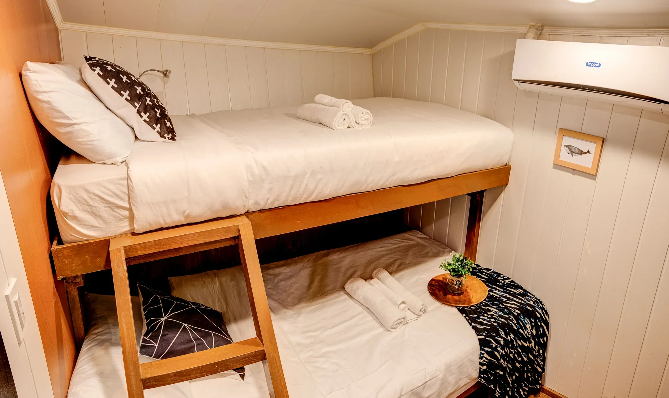 A cozy bedroom with a top bunk bed, white bedding, rolled towels on top, black and white pillows, a small wooden ladder, a wall-mounted air conditioner, a framed fish artwork, and a small table with a plant, blanket, and rolled towel.