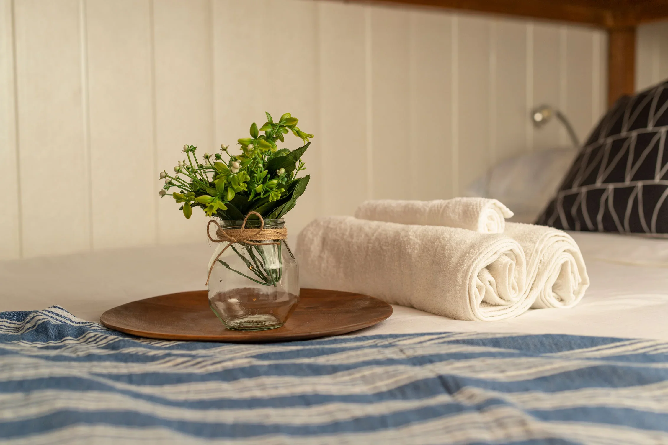 A cozy indoor scene with rolled white towels, a small plant in a jar with twine, and a wooden tray on a bed with a blue and white striped blanket. In the background, part of a black and white patterned pillow and a wooden wall are visible.