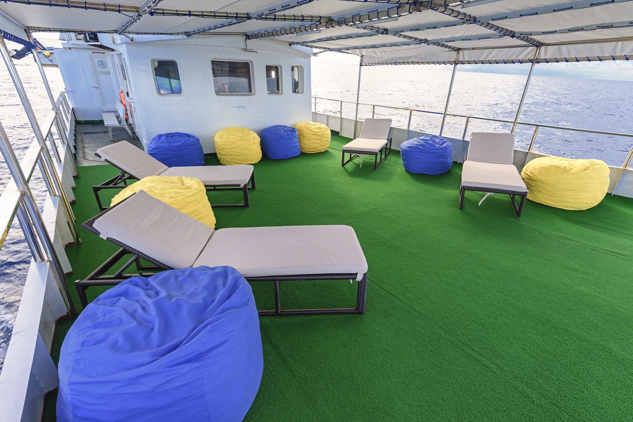 On the deck of a boat, there are lounge chairs, blue and yellow bean bags, and a green carpet. The boat is on open water with a view of the ocean and a cloudy sky in the background.