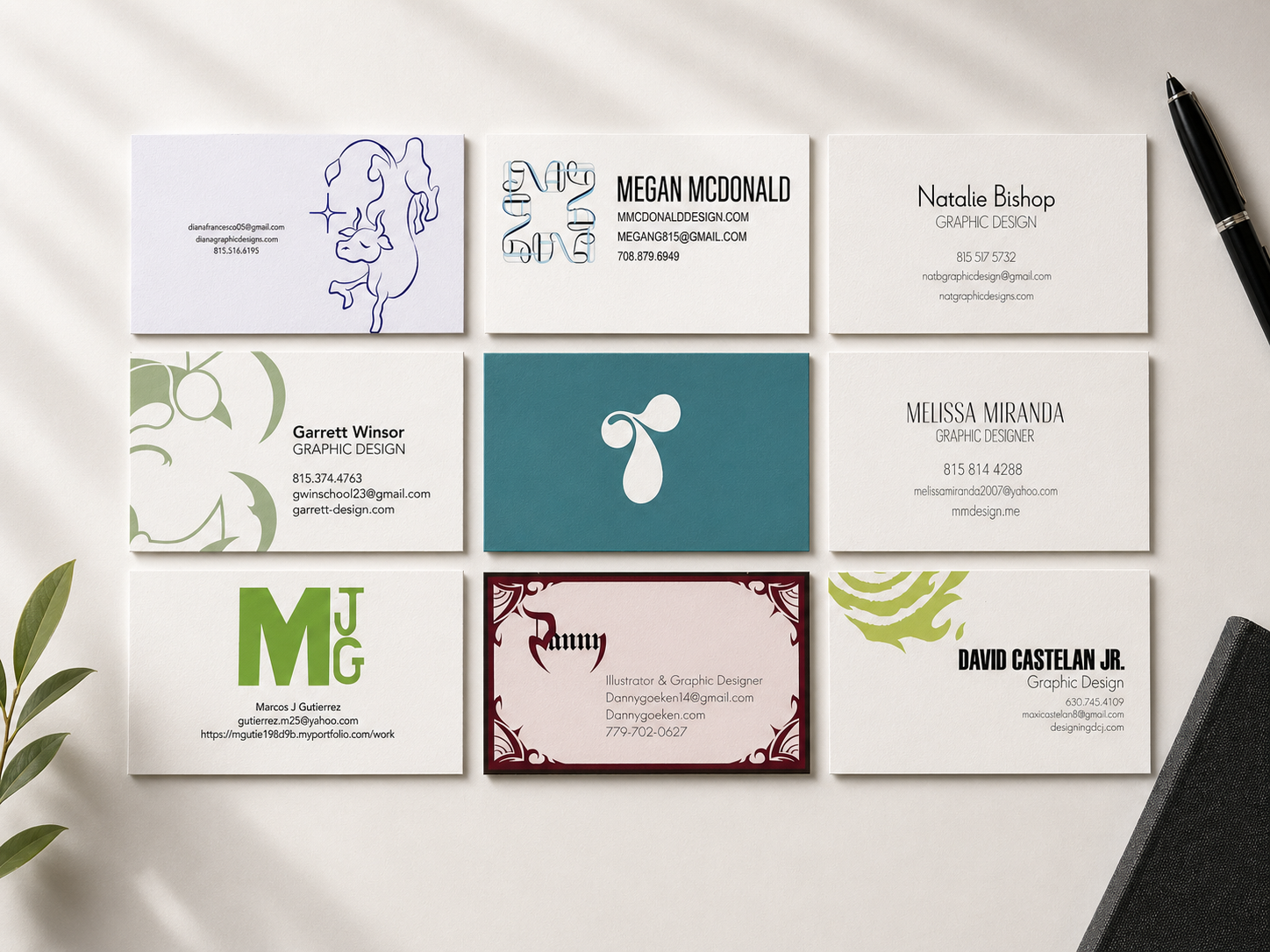 Collection of nine business cards with various designs, logos, and contact information, arranged on a white surface with a black pen to the top right and some green leaves in the bottom left.