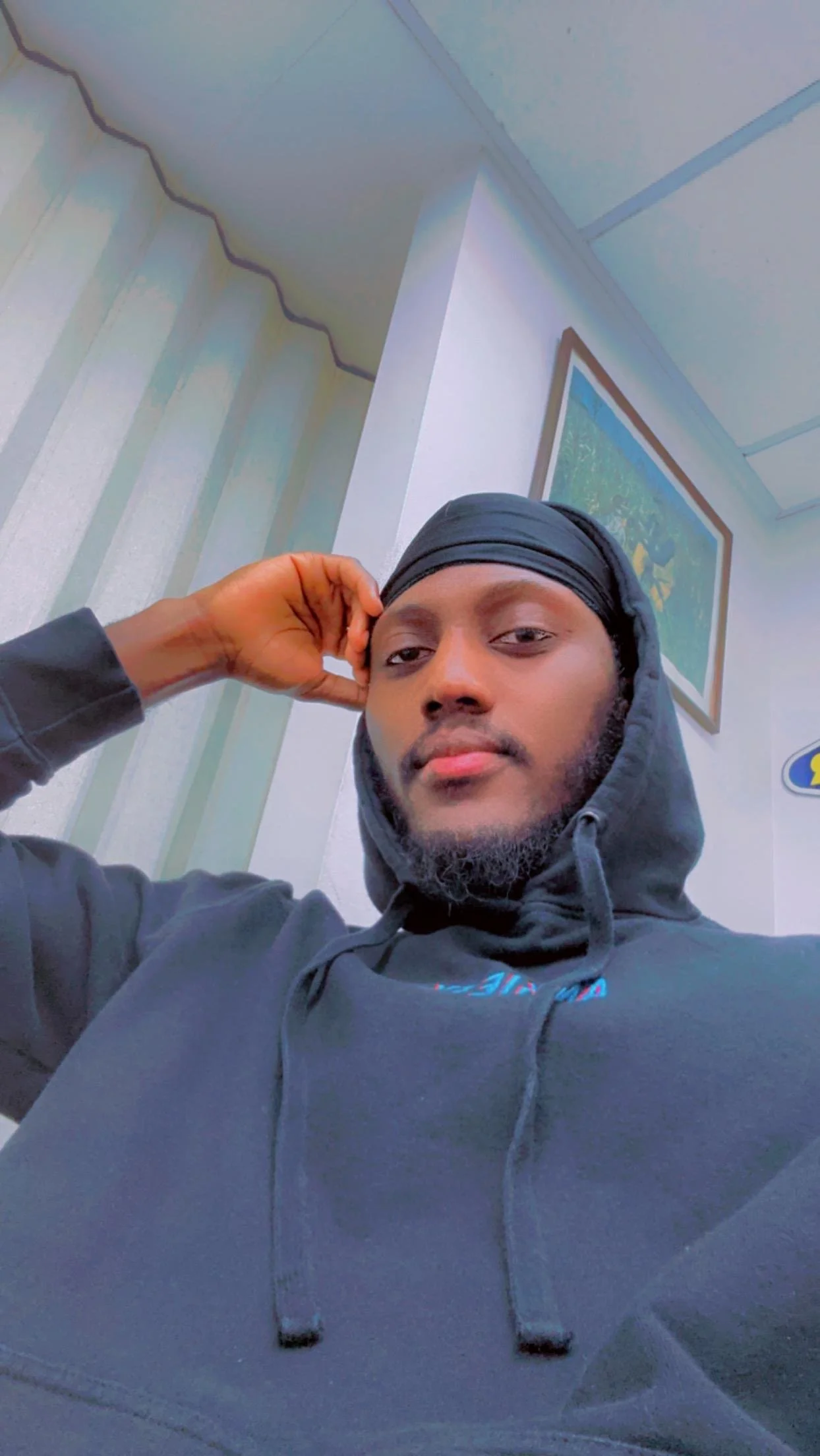 A young man with a beard wearing a gray hoodie and a black durag, resting his head on his hand, taking a selfie indoors with a painting on the wall behind him.